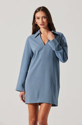 A woman is wearing a loose-fitting, light blue dress with long sleeves. The dress features a pointed collar and a hemline that falls above the knees. The fabric appears to be smooth and slightly textured.